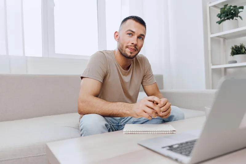 Thoughtful man sitting on a couch, engaging with a laptop, in a modern living room. Warm colors and natural light create an inviting atmosphere for productivity.