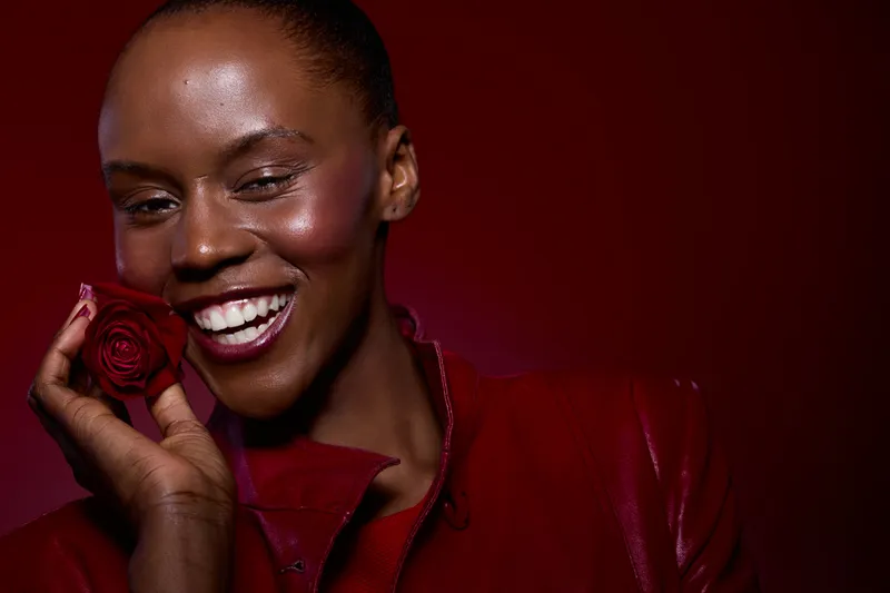 Smiling dark skinned African woman with smooth skin holds red rose near face, wearing red leather jacket on dark red background. Concept for beauty, happiness, natural skin and floral elegance.