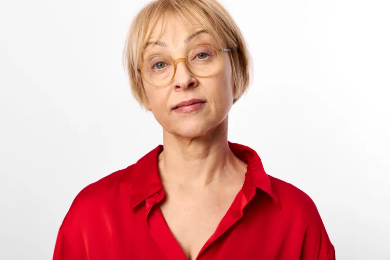 Portrait of senior woman with glasses, blond short hair, wearing red shirt in neutral studio background, calm expression, headshot, soft light.
