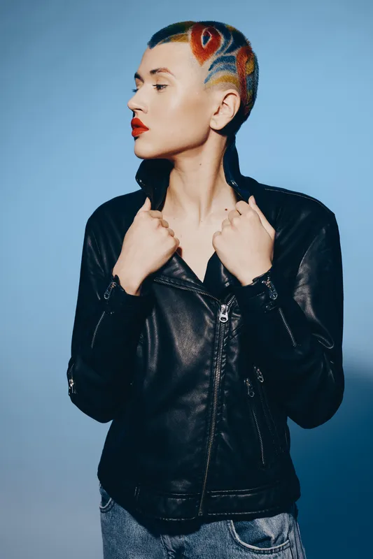Woman with shaved colorful hair wearing black leather jacket and blue jeans stands confidently against blue background expressing modern urban style and individuality.