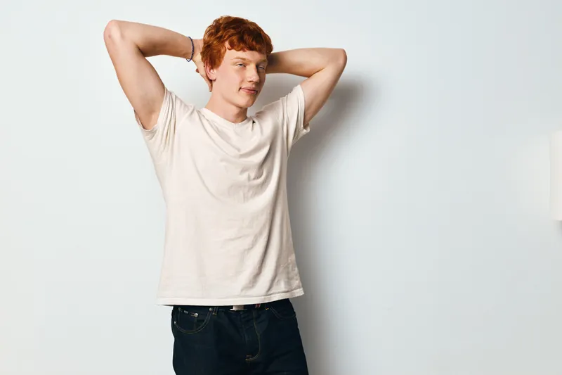 Young man with red hair wearing casual white shirt and dark jeans standing relaxed with hands behind head against plain light wall background indoors.