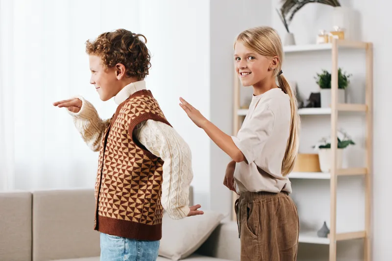 Two children indoors perform playful movement in a cozy living room, a girl and a boy dancing with light from windows, capturing a candid moment of imagination and joy.