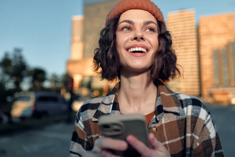 Young woman smile with phone in city at golden hour, candid lifestyle moment in beanie; authenticity and mindful living captured with golden hour glow and emotional storytelling.