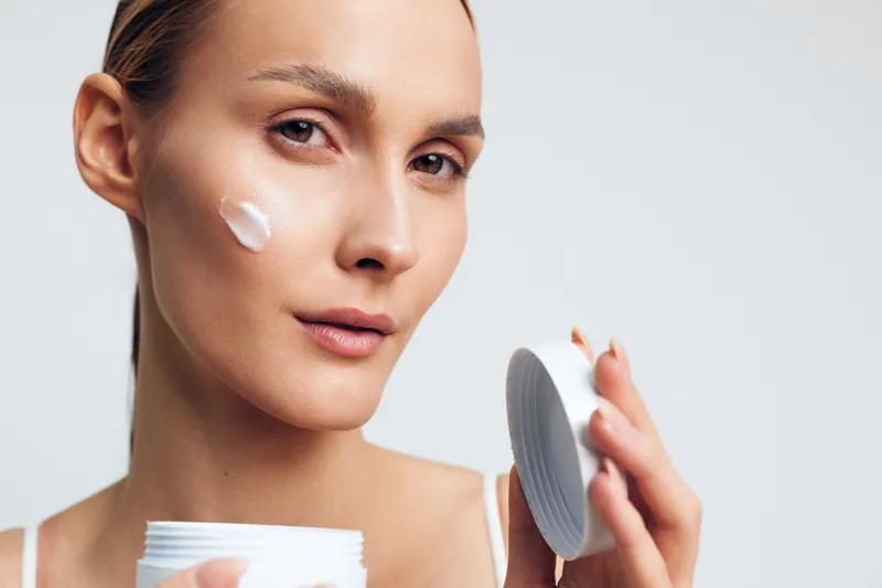 Woman applying face cream in a studio closeup, holding a white jar and smoothing product on skin, skincare beauty selfcare wellness concept, soft light on clean background