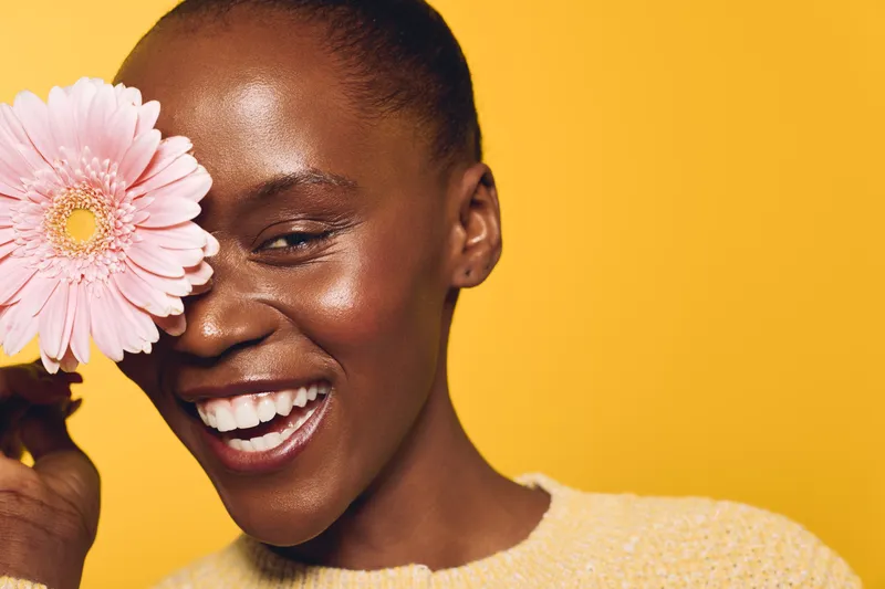 Smiling African woman with dark skin holds pink flower near face on yellow background. Concept of natural beauty, skincare, happiness, and wellness with bright studio lighting.