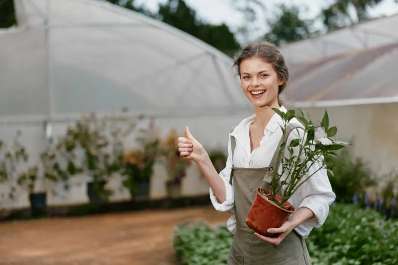 Cheerful young woman in a greenhouse holding a potted plant, dressed in casual gardening attire with a bright smile, symbolizing sustainable living and gardening joy