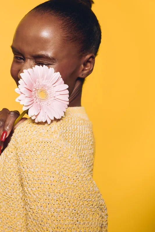 Young black woman with dark skin holds pink flower near face, smiling and winking. She wears yellow knitted sweater on bright yellow background. Concept of beauty, joy, and natural skin care.