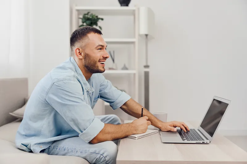 Smiling man working on a laptop at home, showcasing productivity and modern lifestyle in a cozy, minimalist environment with soft natural light.