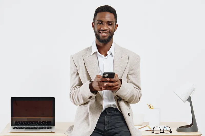 Smiling man using smartphone in bright office space, wearing casual blazer, standing in front of desk with laptop, glasses, and lamp. Office workspace, technology, communication, work from home concep