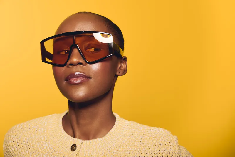 Confident dark skinned African woman with shaved head wearing large orange sunglasses and beige knit sweater. Studio portrait on yellow background. Concept of fashion and style.