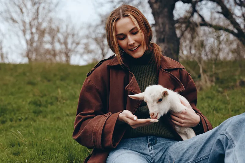 Young woman in brown jacket holds a white baby goat while sitting on grass in a natural outdoor setting during autumn.