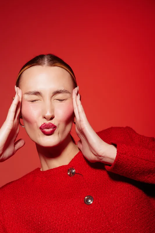 Young woman in red sweater makes playful kiss face with closed eyes and hands on temples. Bright red background and soft lighting create vibrant and cheerful mood in studio portrait.