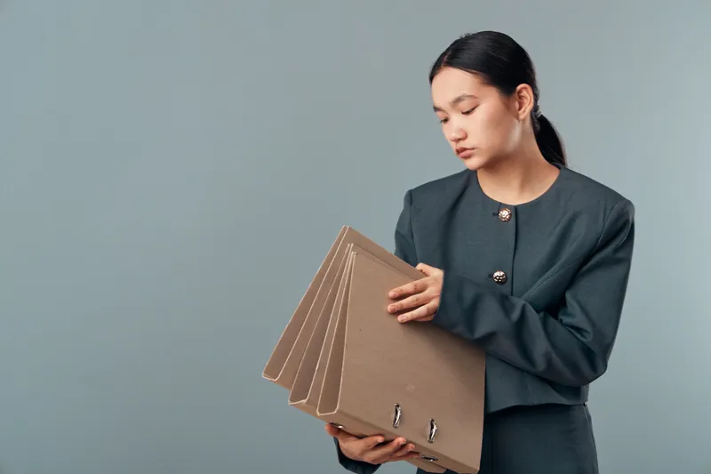 Woman in formal suit holding multiple cardboard folders, office worker organizing documents and files, business preparation and record keeping on gray background.