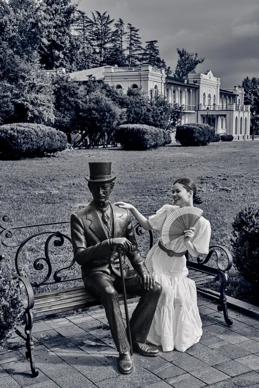 Monochrome scene of a seated couple on a decorative bench with a life size statue, a top hat gentleman and a smiling woman in vintage dress amid a formal garden.