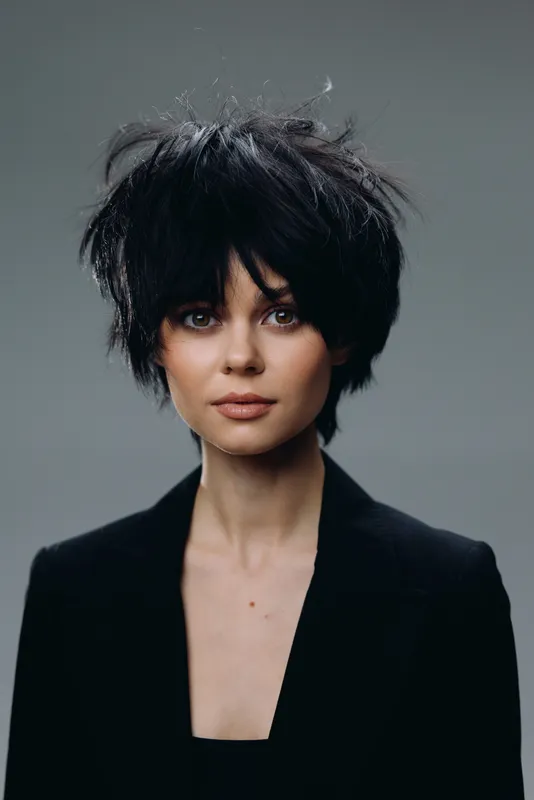 Portrait of a young woman with short black hair wearing a black blazer, expressing calm confidence against a neutral gray background.