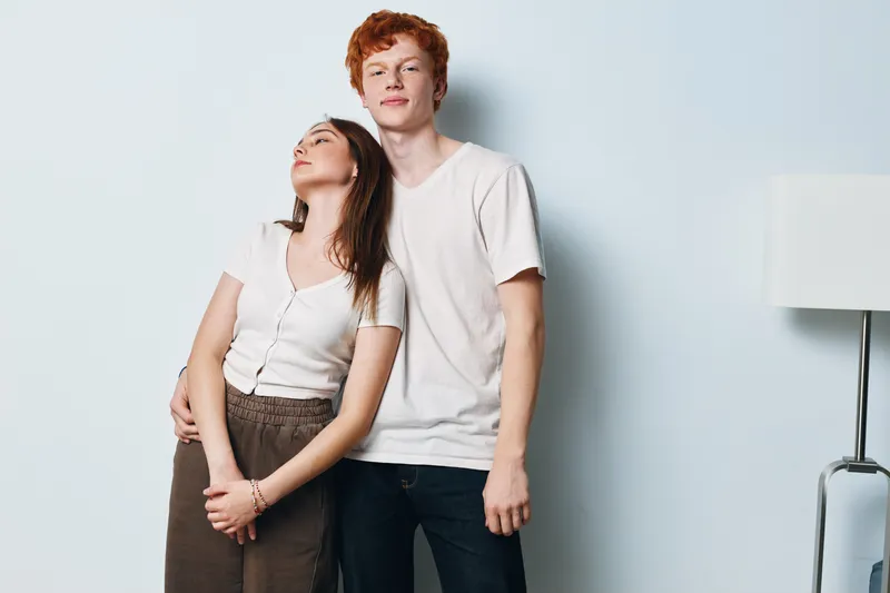 Young man and woman standing together in casual clothes posing near white wall in modern room with lamp. Redhead male and brunette female showing affection and relaxed attitude indoors.
