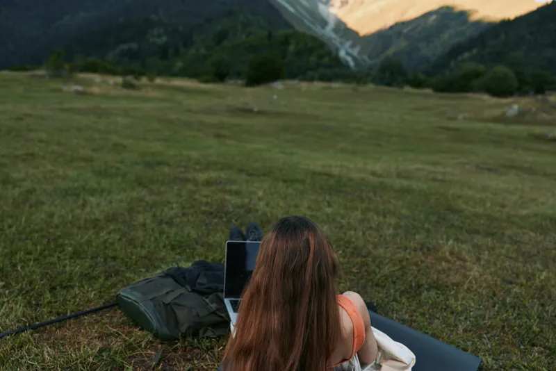 A person sits on a mat in a grassy field with a laptop, enjoying a peaceful outdoor workspace. Natural light and open scenery create a positive, productive remote work atmosphere.