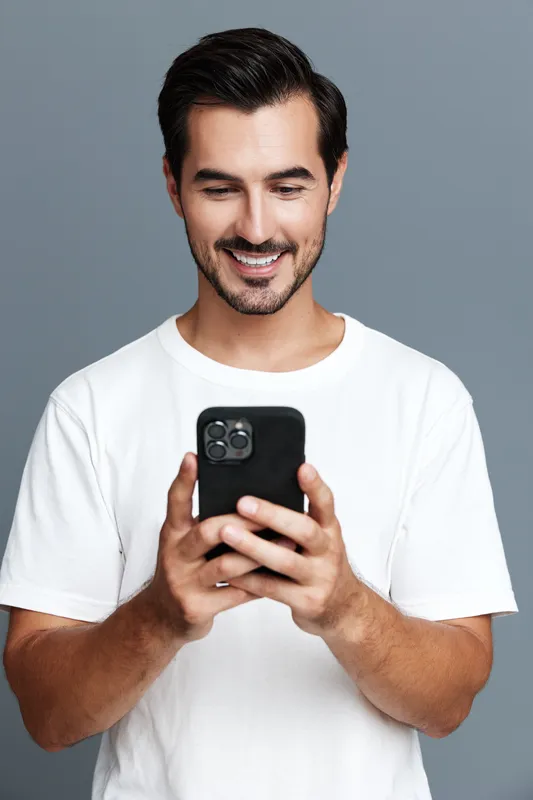 Smiling man wearing a white t-shirt looks at his smartphone with a cheerful expression, isolated on a solid grey background. Modern lifestyle concept.