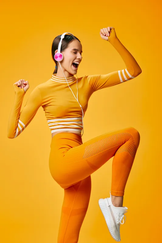 Woman in bright orange athletic outfit dancing and jumping with energetic pose, wearing pink headphones in a vibrant fitness scene. Her athletic demeanor and bold colors convey motivation, movement,
