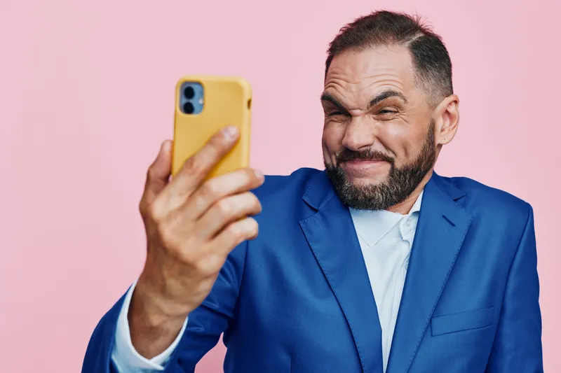 A professional businessman in a blue suit takes a playful selfie against a solid pink background, capturing confident energy and a lighthearted, cheerful mood.