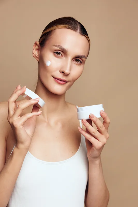 Young woman applies face cream on cheek holding jar and lid with clean skin and natural makeup isolated on beige background, skincare beauty routine concept.