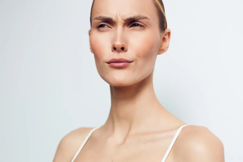 Young woman with serious facial expression looks away with furrowed brows isolated on white background closeup portrait natural skin and minimal makeup visible shoulders.