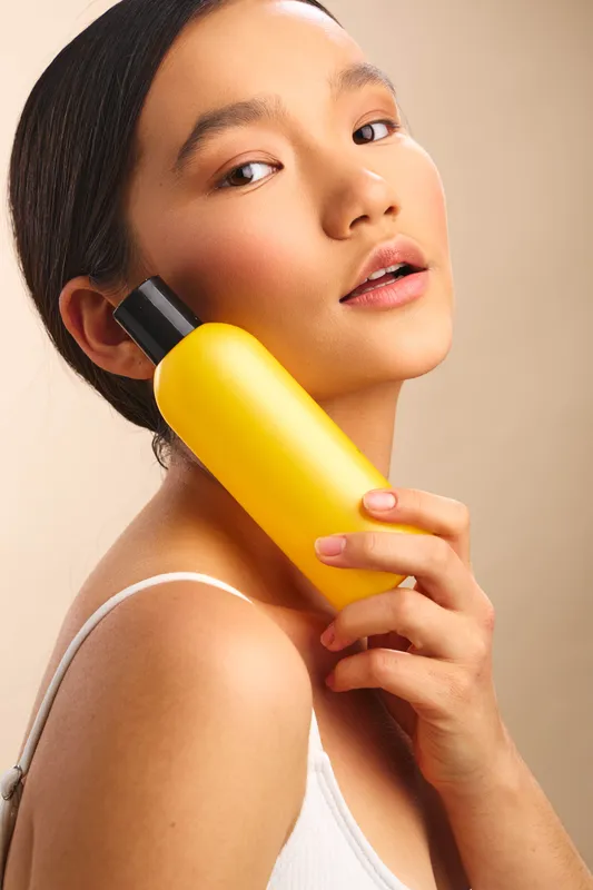 asian woman is holding a bright yellow bottle against her face in a clean studio setting. She gazes softly, conveying freshness and skincare confidence.