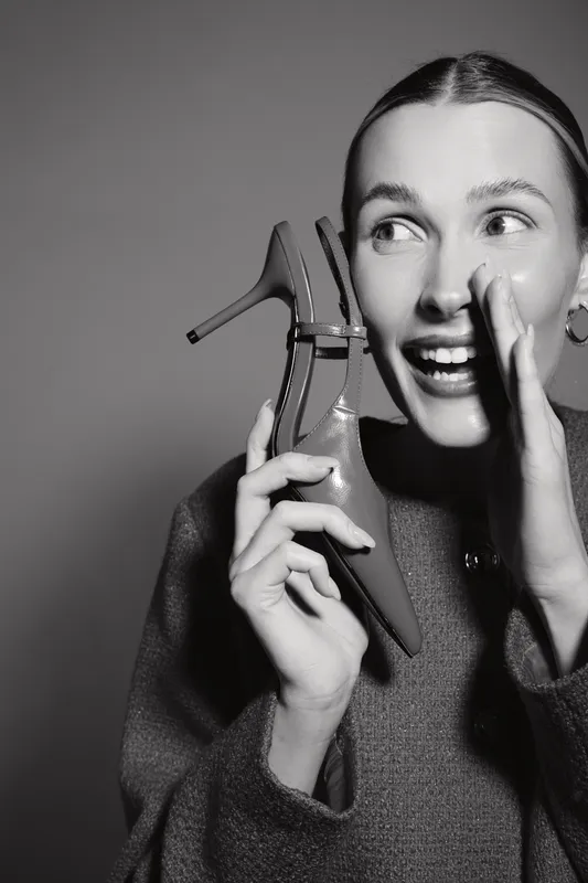 Happy Caucasian woman with light skin holding high heel shoe and whispering with joyful expression in black and white studio portrait.