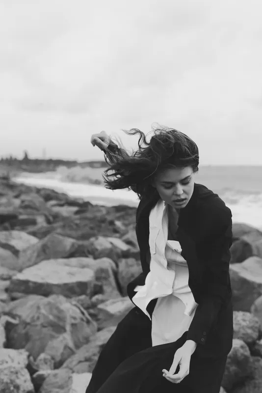 Woman on a rocky shoreline, wind in hair, wearing a shirt and coat, moody monochrome scene by the ocean, dynamic movement and coastal breeze.