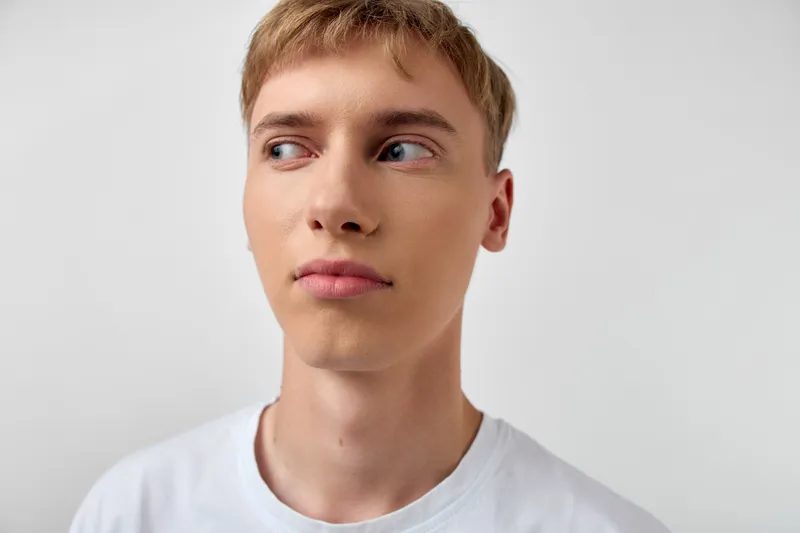 Male with short blond hair looks to the side, neutral expression on a clean white background, close up head and shoulders, simple studio shot emphasizing face and eyes, casual clothing, calm mood,