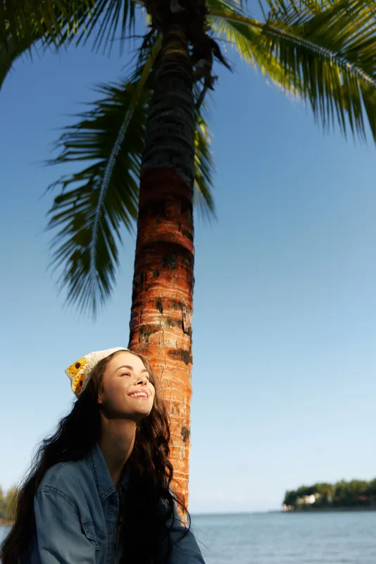 A young woman leans against a tall palm tree beside a calm sea, basking in warm sunlight with a bright smile, wearing a denim jacket and a knit cap, capturing a tropical, carefree moment.
