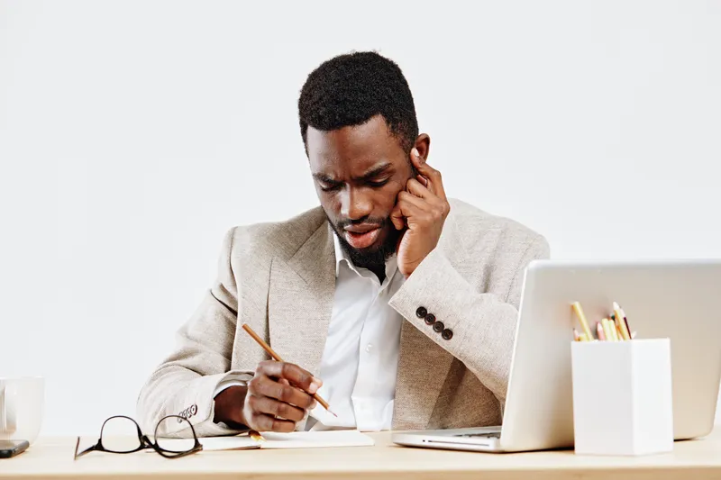 Confused young African American man wearing beige blazer and white shirt sitting at desk with laptop, thinking deeply and holding pen to chin, in bright minimalist office setting. Business concept.