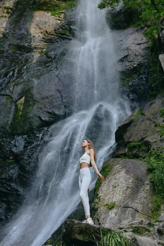 woman standing by waterfall in white activewear, nature, fitness, outdoors, rocks, greenery, serene atmosphere