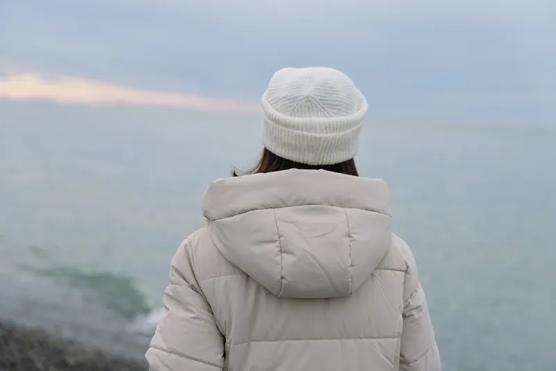 Winter scene with a solitary person viewed from behind, wearing a beige jacket and knit cap by a calm coast and ocean at dusk, creating a peaceful contemplative moment.