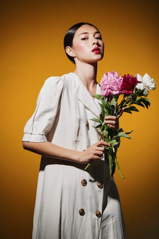 Young woman in minimalist cream dress holding vibrant flowers against a warm yellow background. Concept for beauty creative fashion inspiring elegance and natural style.
