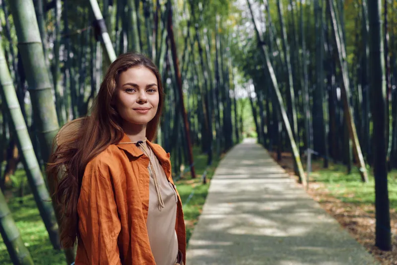 Woman on path in bamboo forest, portrait with warm smile, wearing orange jacket and casual shirt. Nature walkway, peaceful atmosphere and soft sunlight along tree-lined trail.