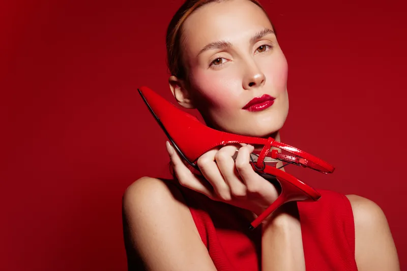 Stylish woman in red dress holds red high heel shoe near face on red background. Fashion concept with bold makeup and elegant pose in studio lighting.