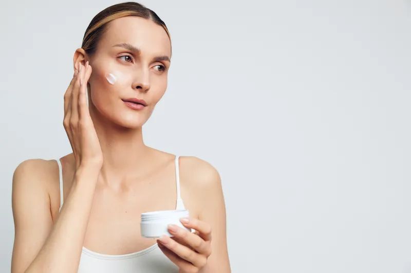 Young woman with calm expression applies moisturizer to her cheek in a clean studio setting, soft light and minimal backdrop, capturing a fresh skincare moment.