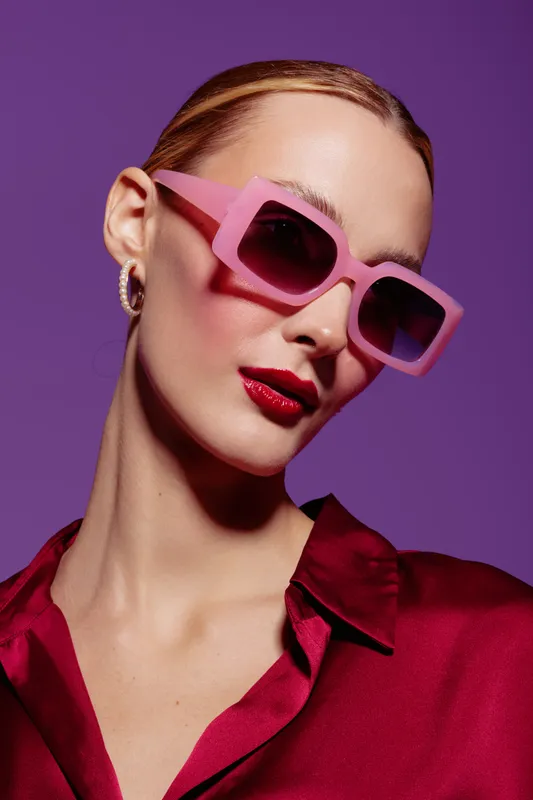 Stylish woman with pink sunglasses and red lipstick poses against purple background wearing a red satin shirt. Fashion portrait with bold colors and modern accessories.