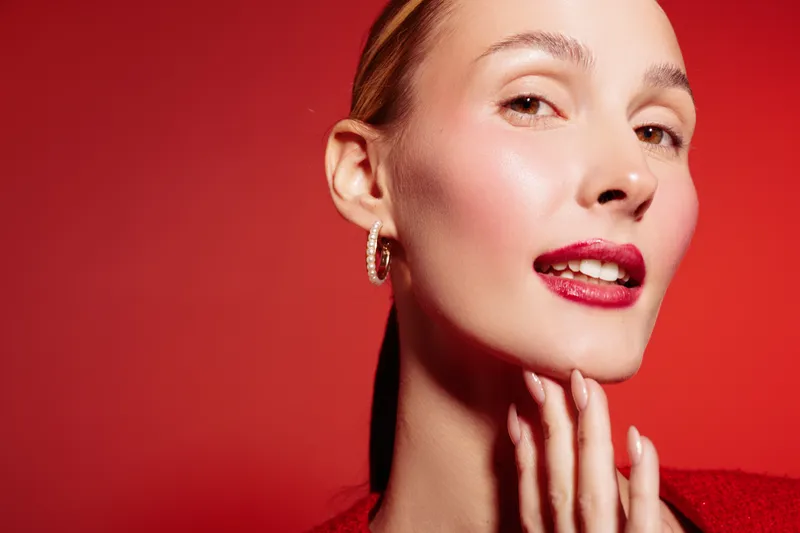 Close-up of a confident woman with red lipstick and hoop earrings posing against a red background. Concept of beauty, fashion, makeup, and self-expression in studio lighting.