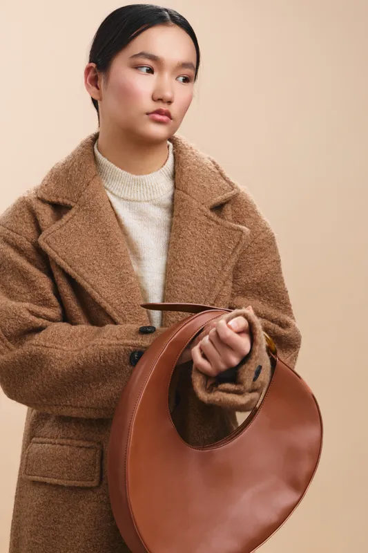 asian woman in a warm brown coat stands in a neutral studio, gazing softly to the side while clutching a round leather bag, conveying calm confidence and modern style.