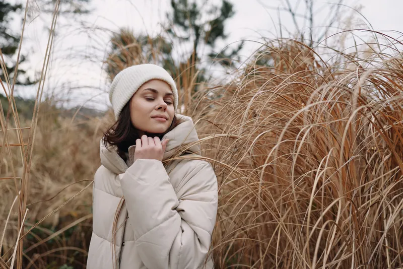 Woman wearing a warm coat and knit hat among tall grasses, a serene winter portrait highlighting outdoor fashion, natural light, calm mood and solitude