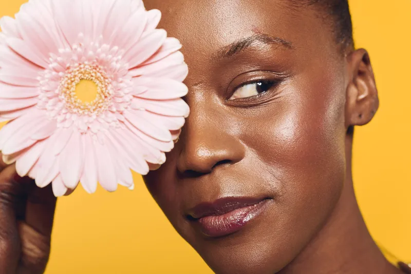 Close-up of a smiling dark skinned African woman with natural makeup holding a pink flower near her face. Bright yellow background creates a vibrant and positive mood for beauty concept.