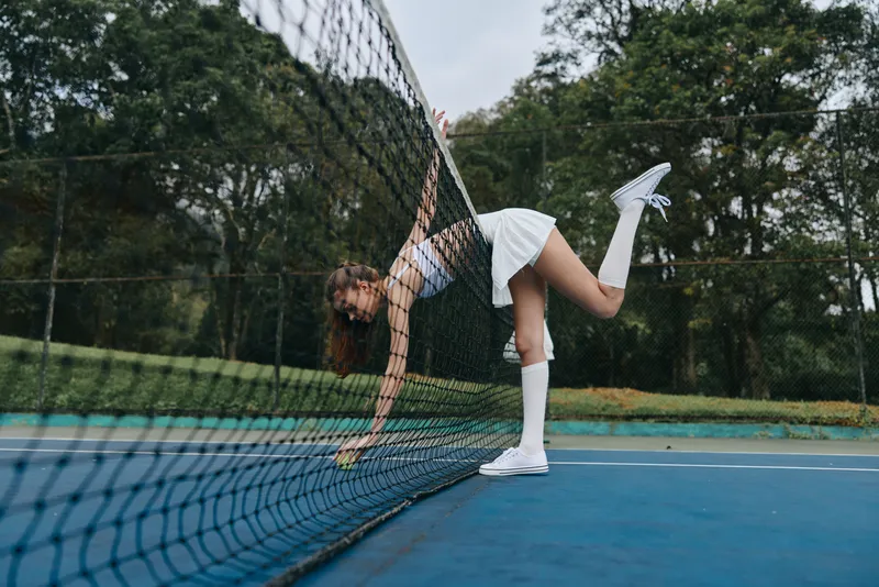 Young female tennis player in a white outfit, joyfully retrieving a ball, showcasing athleticism and energy on a vibrant tennis court
