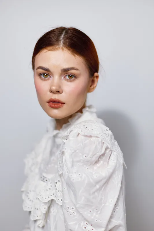 Young woman with red hair in a white ruffled blouse, showcasing an introspective expression against a soft, neutral background