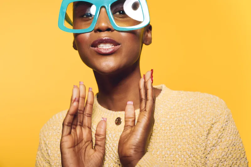 Confident black woman with dark skin wearing oversized blue glasses and a beige sweater poses against yellow background. Concept of fashion, style, and bold accessories.