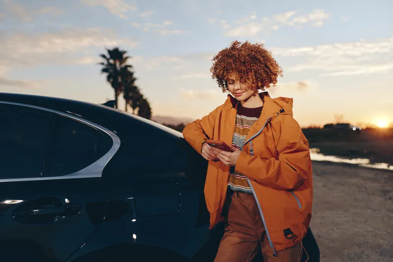Woman lifestyle outdoors at sunset near car, curly hair, orange jacket, holding smartphone, casual travel moment, soft warm evening light, relaxed smile, suburban road background
