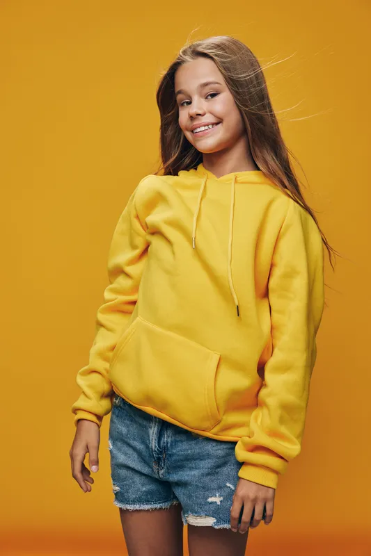 Girl smiling and standing in front of yellow background wearing yellow hoodie and denim shorts. Casual style, happy child, fashion, youth and confident pose.