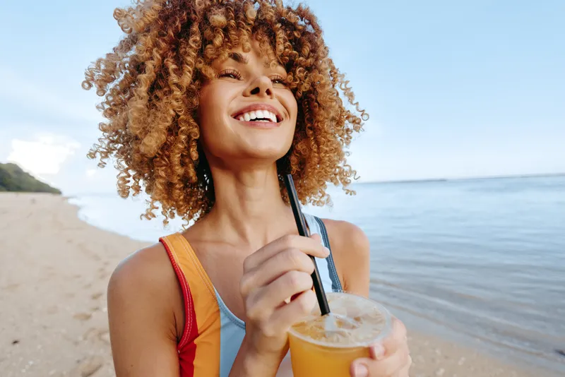 Young woman with curly hair on a sunny beach smiles while sipping an orange juice drink with a straw, enjoying a relaxed moment by the calm sea.