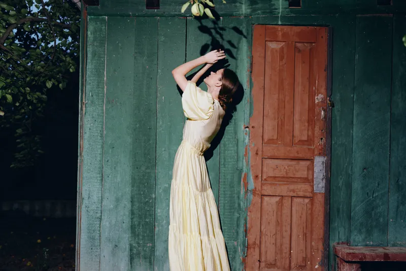 Woman in a yellow dress stands beside a green wall and red door arms raised sunlight warming the scene with vintage charm and a graceful outdoor mood for a timeless portrait